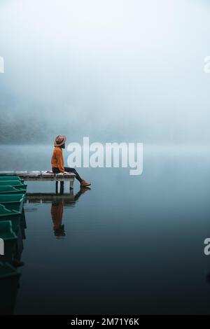 jeune femme à la mode assise sur un quai en bois regardant la vue un matin brumeux Banque D'Images