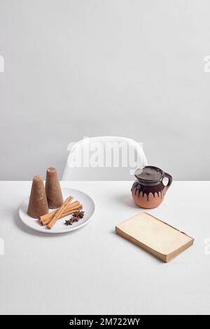 Une table blanche avec une chaise blanche devant un fond blanc, avec du chocolat champurrado dans un pot traditionnel mexicain d'argile, un livre, cannelle, pilon Banque D'Images