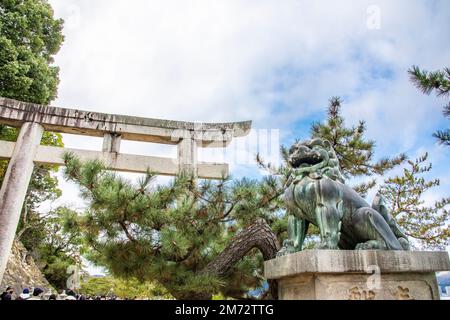 La statue du lion de bronze devant le torii en pierre du sanctuaire d'Itsukushima. Un sanctuaire shinto sur l'île d'Itsukushima (connu sous le nom de Miyajima). Banque D'Images