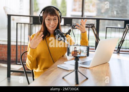 Jeune femme joyeuse podscape influenceur blogueur se gesticulant sur la caméra et souriant tout en faisant le podcast vidéo pour sa chaîne. Une jeune femme diffuse des vidéos en ligne sur un smartphone sur trépied Banque D'Images