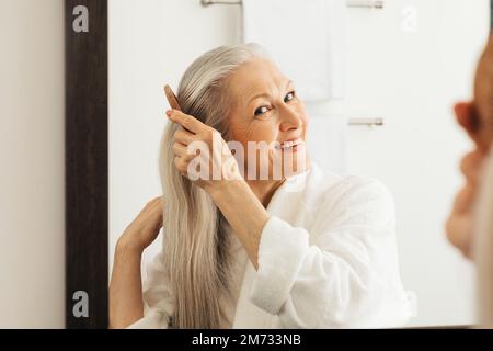 Femme âgée et gaie qui peignait ses cheveux regardant un miroir dans la salle de bains Banque D'Images