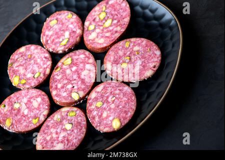 Saucisse traditionnelle de salami fumée avec épices et pistaches sur le noir. Tranches de saucisse de salami sur une assiette noire. Arrière-plan sombre. Banque D'Images