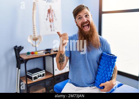 REDHEAD homme avec la longue barbe tenant le rouleau en mousse à la clinique médicale pointant le pouce vers le côté souriant heureux avec la bouche ouverte Banque D'Images