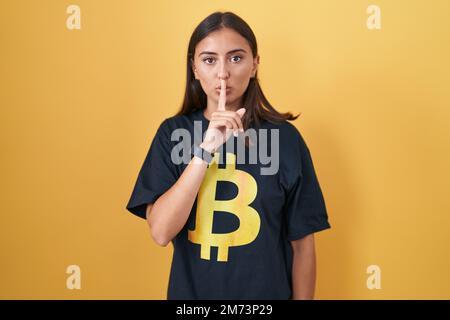 Jeune femme hispanique portant un t-shirt bitcoin demandant à être calme avec le doigt sur les lèvres. silence et concept secret. Banque D'Images