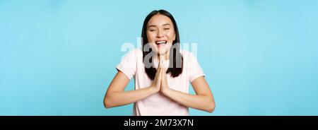 Souriante, la femme asiatique se montre namaste, mendiant geste et rêvant de quelque chose, faisant des rêves, de grands espoirs, se tenant sur fond bleu Banque D'Images