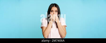 Une femme asiatique choquée couvre la bouche hurle, assourdis, effrayée de sourire, ferme ses lèvres, se tenant sur fond bleu Banque D'Images