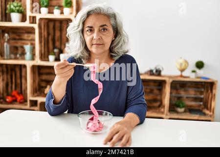 Femme d'âge moyen à cheveux gris mangeant un mètre-ruban assis sur la table à la maison. Banque D'Images