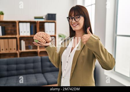 Jeune thérapeute femme travaillant au bureau de thérapie tenant le cerveau souriant heureux et positif, pouce vers le haut faisant excellent et signe d'approbation Banque D'Images