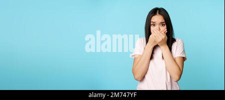 Une femme asiatique choquée couvre la bouche hurle, assourdis, effrayée de sourire, ferme ses lèvres, se tenant sur fond bleu Banque D'Images