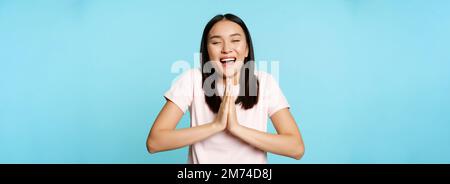 Souriante, la femme asiatique se montre namaste, mendiant geste et rêvant de quelque chose, faisant des rêves, de grands espoirs, se tenant sur fond bleu Banque D'Images