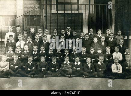 Londres. vers. 1910. Une classe de jeunes élèves et leur enseignant posent pour une photographie de groupe devant leur bâtiment scolaire. Les garçons fréquentent l'école de la rue Gopsall, Hackney. Un garçon tient un tableau affichant le nom de la classe, “Standard IV”. À la suite de la loi de 1876 sur l'éducation, les élèves qui avaient passé l'examen de la norme IV à l'âge de 10 ans et qui détenaient un certificat de fréquentation régulière pendant cinq ans ont droit à une éducation gratuite pendant trois ans. Certains des garçons portent des colliers en lin raides. Banque D'Images