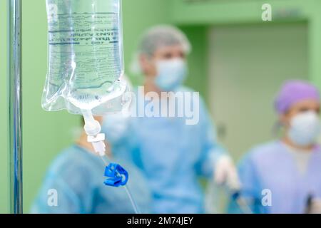 Concentration sélective sur l'emballage contenant une solution saline pour compte-gouttes en salle d'opération. Silhouettes de chirurgiens sur un fond flou. Banque D'Images