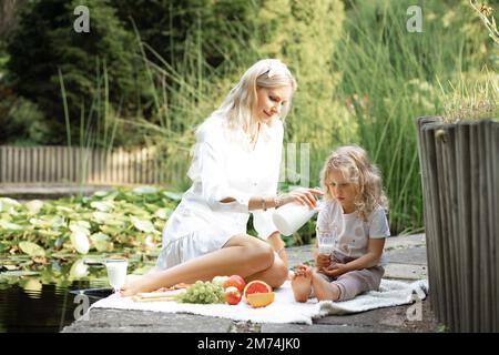 Blond verse du lait dans un verre pour une petite fille dans un parc naturel près d'un lac en plein air. Déjeuner pique-nique et alimentation saine Banque D'Images