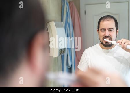 Jeune homme blanc se brossant les dents tout en regardant dans le miroir de sa salle de bains. Banque D'Images