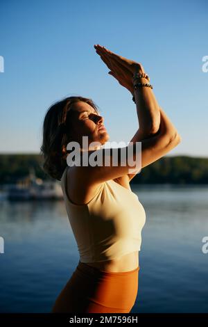 Une femme pratique le yoga près de l'eau dans une posture de yoga d'arbre. Banque D'Images