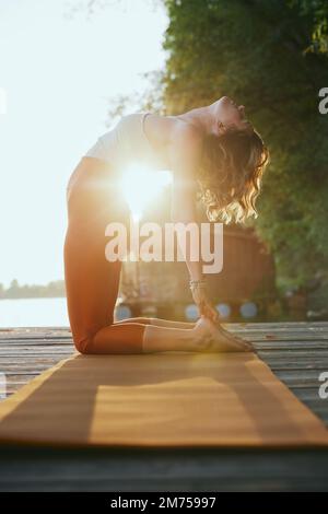 Une femme d'âge moyen en forme pratique le yoga sur le quai au coucher du soleil. Elle est dans la posture du yoga de Camel. Banque D'Images