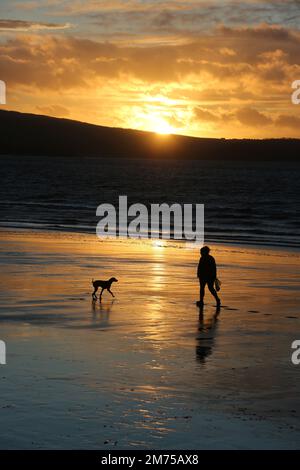 Ayr, Ayrshire, Écosse. UK Jan 2023 .Une figure silhouettée e avec le chien aime une promenade en soirée le long de la plage à Ayr comme l'espace de couchers de soleil pour la copie ou le texte. Image romantique de style de vie Banque D'Images