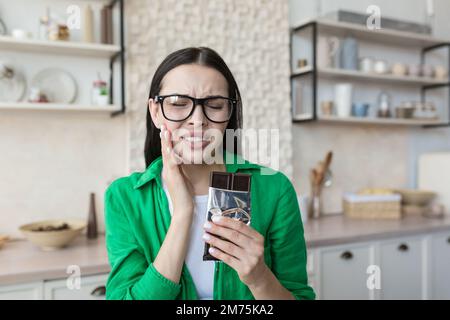 Une jeune femme est assise dans la cuisine à la table à la maison, mangeant une barre de chocolat noir. Tient sa joue, sent un mal de dents fort de salé. Elle grimaca dans la douleur. Banque D'Images