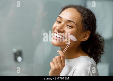 Femme souriante faisant un massage du visage en utilisant un rouleau avec du quartz dans la salle de bains. Concept de cosmétiques naturels Banque D'Images