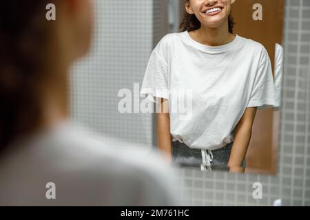 Gros plan d'une femme africaine souriante debout dans la salle de bains et regardant dans le miroir Banque D'Images