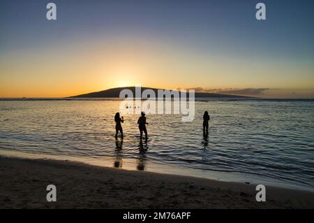 Des jeunes silhouetés et méconnaissables pataugent dans les eaux peu profondes de Baby Beach à Maui avec Lanai au loin. Banque D'Images