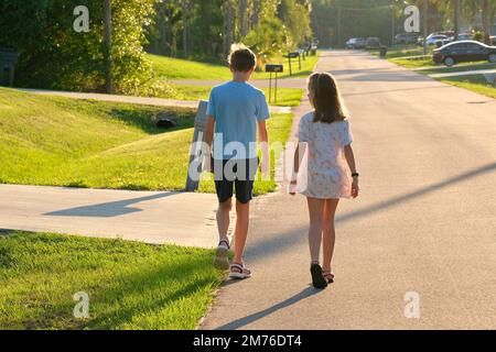 Vue arrière de deux jeunes enfants adolescents, fille et garçon, frère et sœur marchant ensemble dans la rue de banlieue le soir ensoleillé Banque D'Images