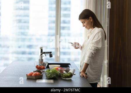 Jeune femme utilisant un smartphone dans la cuisine tout en cuisant de la salade végétarienne Banque D'Images