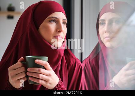 Femme musulmane dans le hijab avec une tasse de café regardant par une fenêtre. Banque D'Images