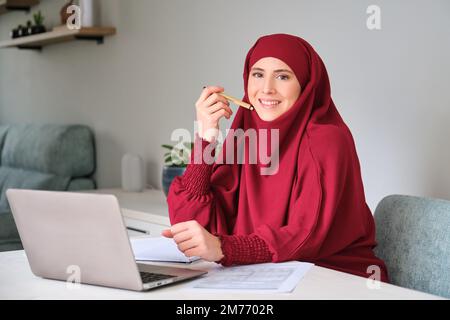 Portrait de la jeune femme musulmane dans le hijab utilisant un ordinateur portable à la maison. Banque D'Images