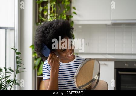 Bonne femme afro-américaine prend soin des cheveux regarde dans le miroir avec le peigne en plastique se trouve dans l'appartement Banque D'Images