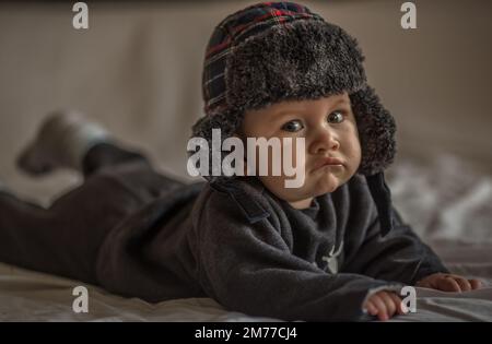 Bébé garçon sur un lit sur son ventre avec la tête vers le haut regardant avec ses grands yeux. Vêtements de biege chauds et molletonnés. Gros plan. Bébé de trois mois Banque D'Images