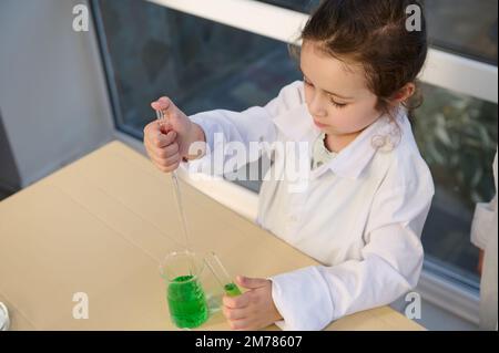 Petite fille en blouse de laboratoire, goutte de liquide chimique dans un bécher, à l'aide d'une pipette graduée en laboratoire de chimie Banque D'Images