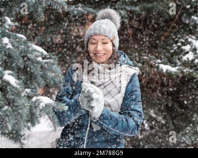 Femme souriante dans le chapeau tricoté de câble joue avec la neige. Amusez-vous dans le parc entre des sapins enneigés. Une femme rit alors qu'elle lance un ballon de neige. Saison froide. Banque D'Images