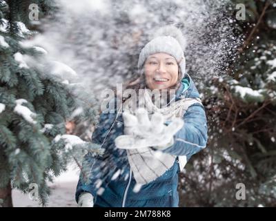 Femme souriante dans le chapeau tricoté de câble joue avec la neige. Amusez-vous dans le parc entre des sapins enneigés. Une femme rit alors qu'elle lance un ballon de neige. Saison froide. Banque D'Images