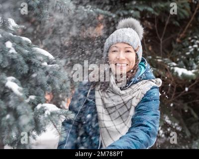 Femme souriante dans le chapeau tricoté de câble joue avec la neige. Amusez-vous dans le parc entre des sapins enneigés. Une femme rit alors qu'elle lance un ballon de neige. Saison froide. Banque D'Images