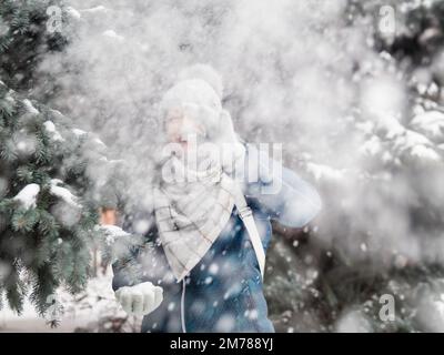 Femme souriante dans le chapeau tricoté de câble joue avec la neige. Amusez-vous dans le parc entre des sapins enneigés. Une femme rit alors qu'elle lance un ballon de neige. Saison froide. Banque D'Images