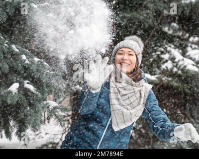 Femme souriante dans le chapeau tricoté de câble joue avec la neige. Amusez-vous dans le parc entre des sapins enneigés. Une femme rit alors qu'elle lance un ballon de neige. Saison froide. S Banque D'Images