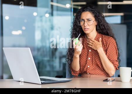 Une jeune femme latino-américaine malade, assise au bureau à une table avec un ordinateur portable, se sent mal. Il tousse, souffle sa gorge avec un aérosol. Tient sa poitrine, il regarde malheureusement la caméra. Banque D'Images