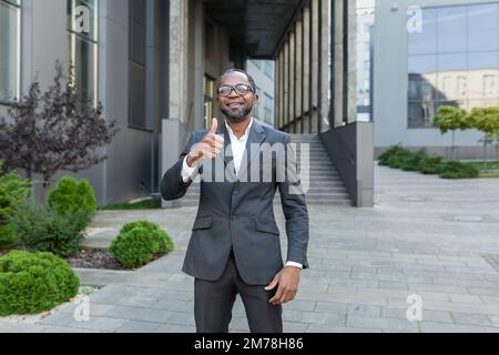 Portrait d'un homme afro-américain réussi. Un homme d'affaires, un avocat se tient devant un bureau dans un costume, regarde la caméra, pointe un super doigt, sourit. Banque D'Images