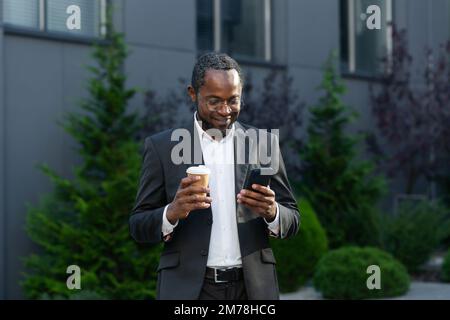 Pause café. Un jeune homme d'affaires afro-américain, indépendant, se trouve dans la rue près du centre de bureaux. Il tient une tasse de papier de café dans ses mains, regarde le téléphone, sourit. Banque D'Images