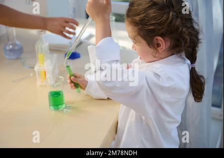Vue latérale d'un enfant intelligent à l'aide d'une pipette graduée et de verres dosing, en faisant des expériences dans un laboratoire de chimie scolaire Banque D'Images