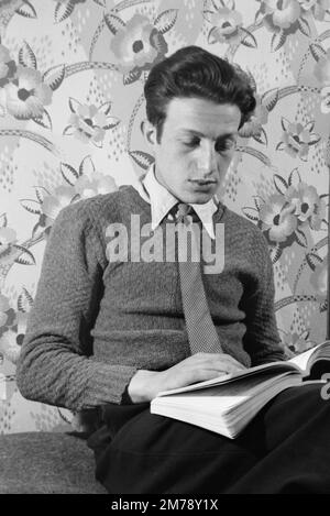 1940s Portrait d'un jeune homme français portant 1940s vêtements et lisant un livre de papier sur un fond de 1940s papier peint floral France c1945. Photographie noir et blanc ou monochrome vintage. Banque D'Images