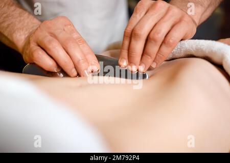 Vue rapprochée des mains de l'homme qui massent l'estomac d'une femme à l'aide de l'outil IASTM pour gratter les muscles dans le spa ou la clinique de réadaptation Banque D'Images