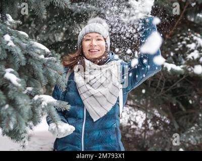 Femme souriante dans le chapeau tricoté de câble joue avec la neige. Amusez-vous dans le parc entre des sapins enneigés. Une femme rit alors qu'elle lance un ballon de neige. Saison froide. Banque D'Images