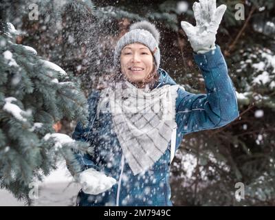 Femme souriante dans le chapeau tricoté de câble joue avec la neige. Amusez-vous dans le parc entre des sapins enneigés. Une femme rit alors qu'elle lance un ballon de neige. Saison froide. Banque D'Images