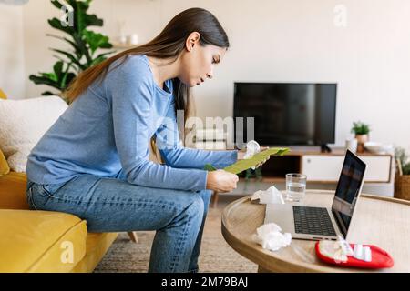 Jeune femme lisant les instructions de traitement tout en ayant un appel vidéo avec un médecin Banque D'Images