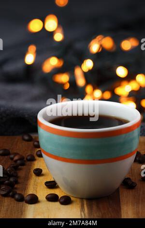 Image du mode de vie : une tasse de café noir coloré est placée sur une surface en bois avec des grains de café au premier plan et des lumières bokeh en arrière-plan. Banque D'Images