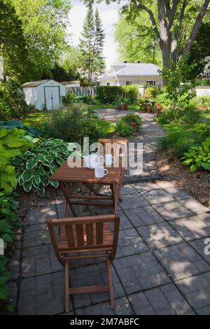 Table et chaises en bois brun sur le patio de carreaux de pavage de ciment gris à côté des frontières avec des plantes Hosta dans le jardin paysager de la cour arrière au printemps. Banque D'Images