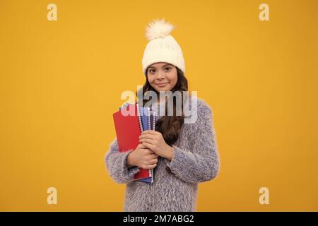 Adolescent dans un chandail chaud et un chapeau tenir des livres et un sac à dos isolés sur fond jaune. Bonne écolière, émotions positives et souriantes. Banque D'Images