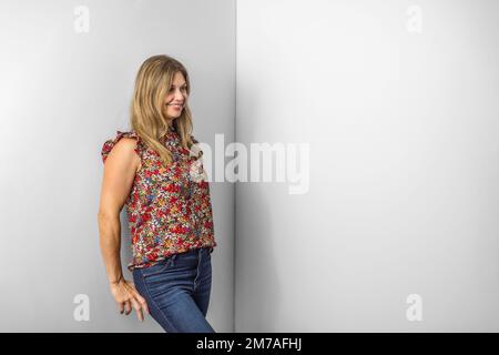 une belle femme caucasienne dans ses années quarante dans un haut fleuri décontracté et jeans en denim debout contre un mur blanc avec espace de copie Banque D'Images